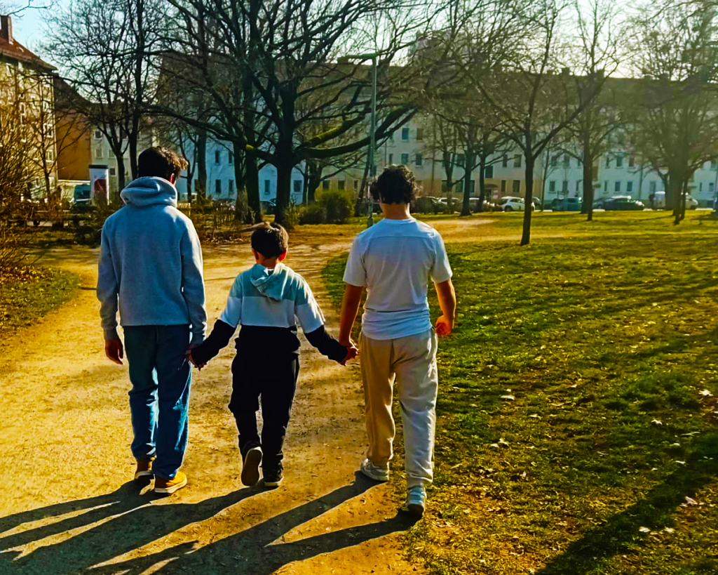 Drei Brüder gehen gemeinsam auf einem Weg im Park. Der Kleine in der Mitte, hält die Hände seiner Brüder links und rechts. Die Bäume im Hintergrund haben keine Blätter, was auf Herbst oder Winter hindeutet.