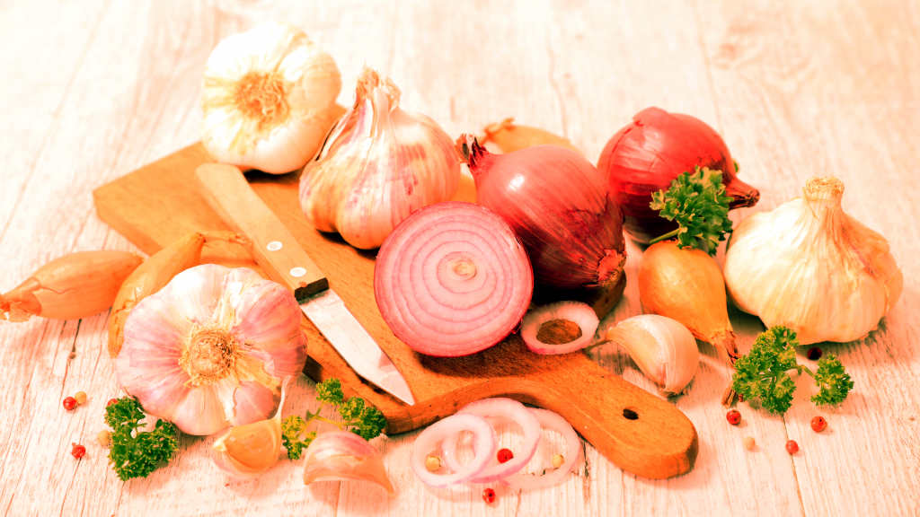 Auf einem hölzernen Schneidebrett sind verschiedene frische Zutaten angeordnet: Knoblauchknollen, rote Zwiebeln, Schalotten und eine aufgeschnittene rote Zwiebel. Ein Messer liegt bereit, während Petersilienzweige und Pfefferkörner die Szene abrunden.