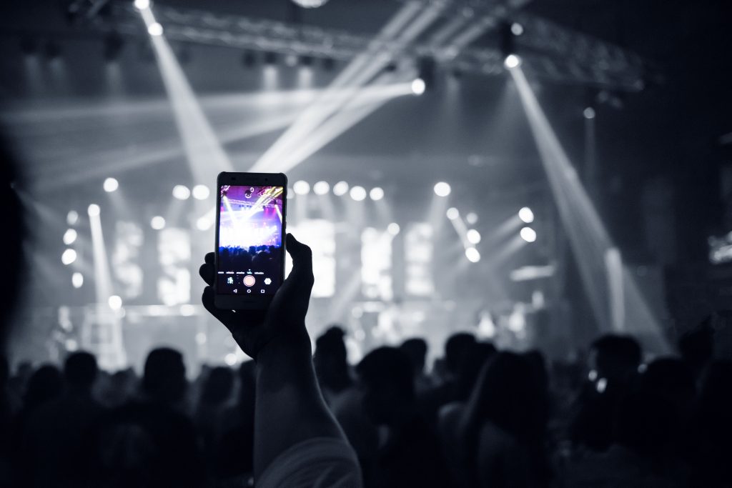 Eine Person hält ein Smartphone hoch und filmt ein Konzert oder eine Veranstaltung. Im Hintergrund sind viele Menschen und helle Bühnenlichter zu sehen.