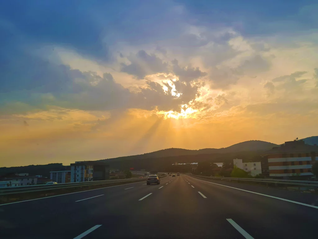 „Eine Autobahn erstreckt sich unter einem beeindruckenden Himmel, während die Sonne goldenes Licht auf die Straße wirft. Autos sind unterwegs, und Gebäude säumen die Fahrbahn.